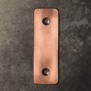 May include: A rectangular copper-toned metal plate with a brushed finish and rounded corners. The plate features two circular, countersunk screw holes positioned vertically along the center line, one near the top and one near the bottom edge.