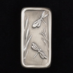 May include: A rectangular, silver-toned metal object with a raised relief design of two dragonflies and aquatic plants. The dragonflies have detailed wings, and the plants include reeds and delicate branches.