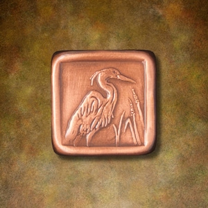 May include: A square copper-toned tile featuring a raised relief design of a heron standing among tall reeds. The tile has a smooth, rounded border and a metallic finish with darker shading in the recessed areas to highlight the bird's form.