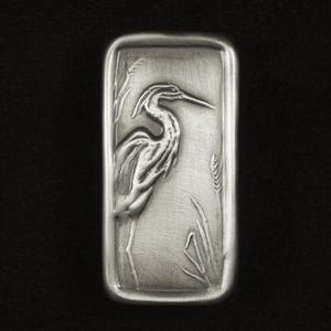 May include: A rectangular, silver-toned metal bar featuring a detailed relief of a heron standing among reeds and wheat. The heron is depicted with its long beak and slender legs. The background is a brushed metal texture.