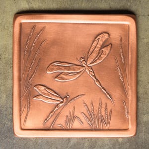May include: A square copper tile featuring a relief design of two dragonflies hovering over stalks of wheat. The metal surface has a warm, metallic reddish-brown finish with embossed details showing the wings, bodies, and textured grass blades.