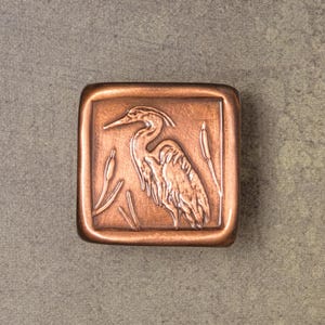 May include: A square copper-toned metal tile featuring a raised relief design of a heron standing among cattails. The piece has a smooth, rounded border and a warm, metallic finish with subtle variations in color across the textured surface.