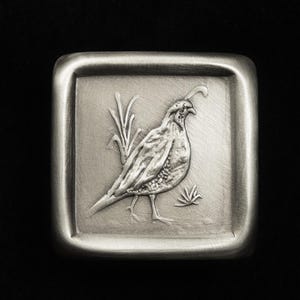 May include: A square, silver-toned metal object with a detailed relief of a quail and foliage. The bird is depicted in profile, with textured feathers and a distinctive head plume. The background has a brushed metal finish.