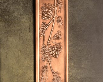 May include: A rectangular copper-toned tray featuring an embossed design of a pine branch with two pinecones. The metal surface has a brushed finish with raised details depicting the needles and texture of the cones centered on the tray.