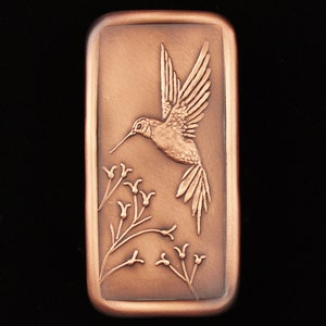 May include: A copper-colored rectangular plaque with an embossed image of a hummingbird in flight, hovering over a branch of small flowers.