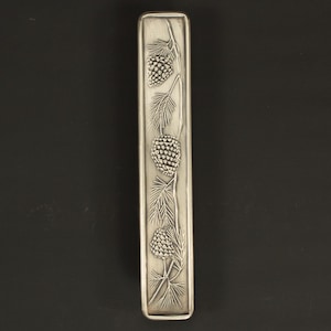 May include: A rectangular, silver-toned metal object with an embossed design of pine cones and needles. The design is centered on the object, with three pine cones visible. The object has a smooth, polished finish.