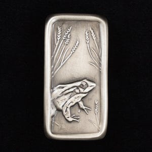 May include: A rectangular silver metal bar featuring a detailed relief of a frog and wheat stalks. The frog is centered, with wheat stalks on either side. The bar has a slightly rounded edge and is set against a black background.