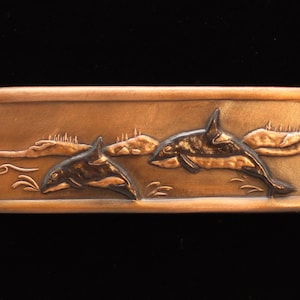 May include: A rectangular copper-colored metal plaque with a raised design of two orcas jumping out of the water. The orcas are black and the water is a lighter copper color.