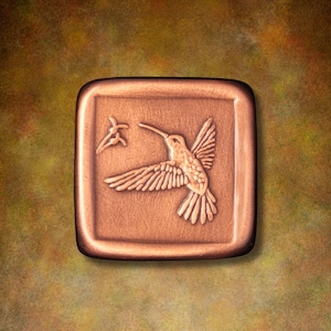 May include: A square copper-toned tile featuring a raised relief design of a hummingbird in flight with wings spread wide. To the left of the bird is a small, stylized floral element. The tile has rounded corners and a smooth, metallic finish.