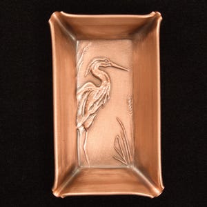 May include: A rectangular copper-colored tray featuring a raised relief design of a heron standing among tall grasses. The tray has a metallic finish with a brushed texture and slightly curved, flared edges, creating a shallow basin shape.