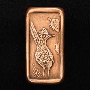 May include: A rectangular copper-colored metal bar with a detailed relief of a roadrunner bird, cacti, and grass. The roadrunner is facing left, with its tail feathers and crest prominently displayed. The bar has a slightly rounded edge.