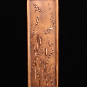 May include: A rectangular copper plaque with an embossed design of cattails and dragonflies. The plaque has a rustic, aged look.