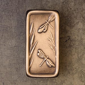 May include: A rectangular bronze-toned metal tray featuring a raised relief design of two dragonflies, cattails, and reeds. The surface has a textured, weathered finish with a prominent border framing the central nature-inspired artwork.