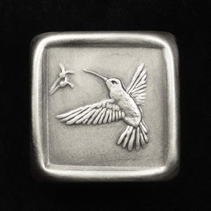 May include: A square, silver-toned metal object with a raised relief design of a hummingbird in flight. The bird is depicted with detailed wings and a long beak, facing a small, abstract floral element. The edges of the object are rounded.