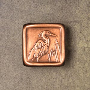 May include: A square-shaped copper-toned tile featuring a raised relief design of a heron standing among tall reeds. The object has a smooth, rounded border and a metallic finish with darker shading in the recessed areas of the embossed bird image.