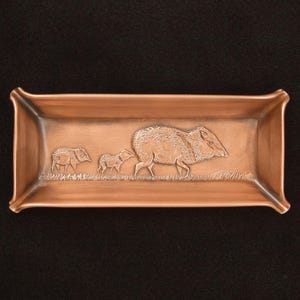 May include: A rectangular copper tray featuring a relief design of a large wild boar followed by two smaller piglets walking across a grassy field. The tray has slightly flared corners and a smooth, metallic finish with warm reddish-brown tones.