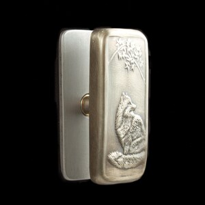 May include: A silver-colored rectangular lighter with a detailed relief of a fox sitting beneath a tree. The lighter is open, revealing the interior and a gold-colored wheel.