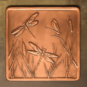 May include: A square copper wall plaque featuring a relief design of three dragonflies flying among cattails and tall grasses. The metal surface has a warm, metallic finish with embossed details of the insects and plants, and a signature in the corner.
