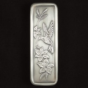 May include: A rectangular, silver-toned metal object with an engraved design of a hummingbird in flight, flowers, and foliage. The hummingbird is depicted with detailed wings and a long beak, surrounded by stylized floral elements.