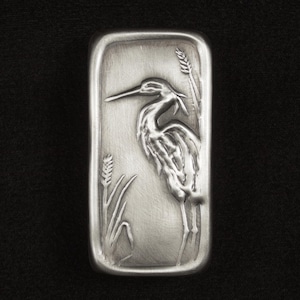 May include: A rectangular, silver-toned metal bar featuring a detailed relief of a heron standing amidst reeds. The bird has a long beak and is surrounded by stylized plant life. The background is a solid black.