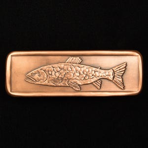 May include: A copper-colored rectangular belt buckle featuring a detailed relief of a fish. The fish is depicted in profile, with scales, fins, and a tail. The buckle has a raised border and a polished finish.