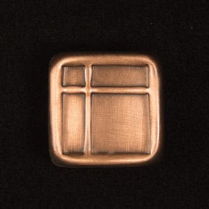 May include: A square, copper-colored metal button with a grid-like design. The button has a slightly rounded edge and a textured surface. The button is set against a black background.