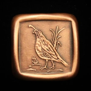 May include: A copper-colored square button with a raised image of a quail standing in a field of grass.
