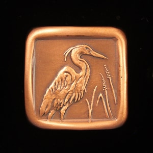 May include: A copper-colored square plaque with a raised image of a heron standing in tall grass.