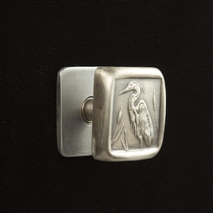 May include: A silver-toned door handle with a square face featuring a detailed relief of a heron standing among reeds. The handle has a rectangular backplate and a curved, cylindrical grip.