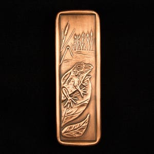 May include: A copper-colored rectangular pendant featuring a detailed relief of a frog on a leaf, with cattails and water in the background. The pendant has a polished finish and is set against a black backdrop.