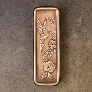 May include: A rectangular bronze-toned metal plate featuring an embossed design of a hummingbird in flight hovering near a flowering branch with two open blossoms and several leaves. The plate has a raised border and a smooth, metallic finish.