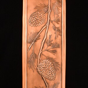 May include: Copper-colored rectangular panel with an embossed design of pine cones and needles. The detailed relief work depicts two pine cones on a branch with surrounding foliage. The background is a solid black.