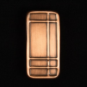 May include: A rectangular, copper-colored metal object with a series of rectangular and square indentations. The object has a slightly rounded top and bottom, and is set against a black background.
