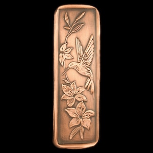 May include: A copper-colored rectangular plaque with an embossed design of a hummingbird in flight, surrounded by flowers. The hummingbird is facing left and has its beak extended towards a flower.