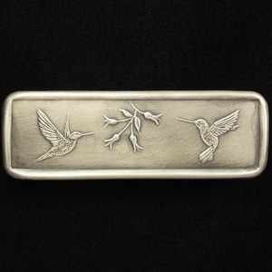 May include: A rectangular, silver-toned metal belt buckle featuring two detailed hummingbird designs flanking a floral sprig. The hummingbirds are depicted in mid-flight, with outstretched wings. The buckle has a slightly textured surface.