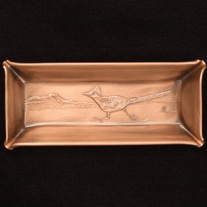 May include: A rectangular copper tray with a raised relief design featuring a roadrunner bird in a desert landscape. The tray has a metallic finish with a warm reddish-brown tone and slightly flared edges, showcasing a detailed embossed scene.