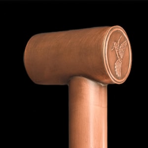 May include: A copper-colored cabinet pull with a cylindrical handle and a circular end. The end features a detailed relief of a hummingbird in flight. The pull has a T-shape design and is set against a black background.