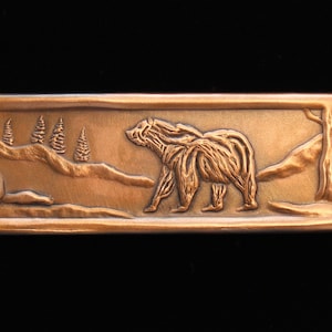 May include: A rectangular copper plaque with a detailed relief sculpture of a bear walking through a forest scene. The bear is facing right and has a textured fur pattern. The background features a mountain range, trees, and a small rock.