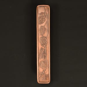 May include: A copper-colored rectangular object with a detailed relief of pine cones and needles. The design is centered on the object, with three pine cones visible. The background is a solid black.