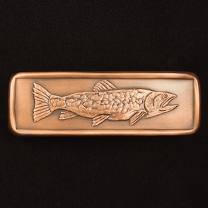 May include: A copper-colored rectangular belt buckle featuring a detailed relief of a fish. The fish is oriented horizontally, with scales and fins clearly visible. The buckle has a slightly rounded frame and a polished finish.