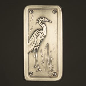 May include: A rectangular, brushed metal plaque featuring a detailed relief of a heron standing among reeds. The heron is depicted with intricate feather detailing. The plaque is secured with four screws, and the artist's signature is visible.