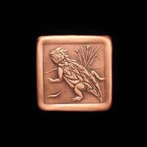 May include: A copper-colored metal square with a raised design of a horned lizard with a spiky back, walking on a textured surface with a plant in the background.