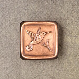 May include: A square copper-toned metal tile featuring a raised relief design of a hummingbird in flight. The bird is positioned on the left, with a small floral sprig depicted on the right. The surface has a brushed, metallic finish.