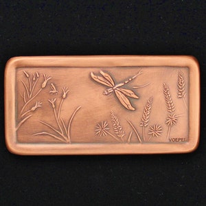 May include: A rectangular copper plaque with an embossed design of a dragonfly flying over tall grass. The plaque is signed "VOEPEL" in the lower right corner.