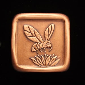 May include: A copper-colored square plaque with a raised design of a bee pollinating a flower. The bee is detailed with wings, legs, and a body. The flower is stylized with petals and a center.