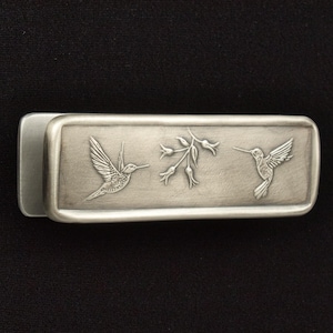 May include: A silver-toned metal money clip featuring a detailed hummingbird design. The rectangular clip has two hummingbirds and a floral element engraved on the front. The clip is shown against a black background.