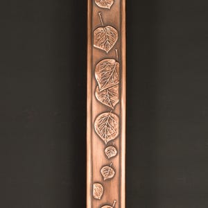 May include: A copper-colored, rectangular decorative element with a series of embossed leaf designs. The leaves vary in size and are arranged vertically along the length of the piece. The background is a solid black.