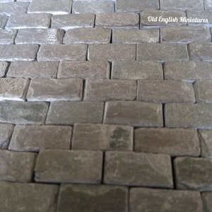 May include: A close-up of a brick walkway made of small, rectangular, gray stones. The stones are arranged in a staggered pattern, creating a textured surface. The text "Old English Miniatures" is visible in the upper right corner of the image.