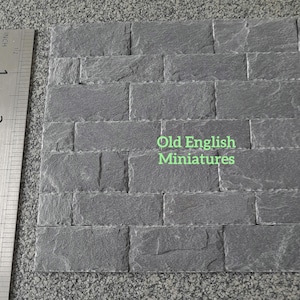 May include: A sample of grey miniature brick veneer, approximately 10 cm by 10 cm, with a ruler for scale. The brick has a rough, textured surface. The text "Old English Miniatures" is visible on the brick. Ideal for dollhouses and model building.