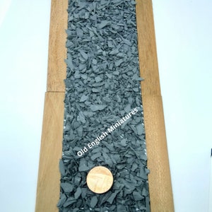May include: A close-up of a bag of gray slate chips for model railroading or miniature landscaping. The bag is next to two wooden planks. A British one-pound coin is included for scale.
